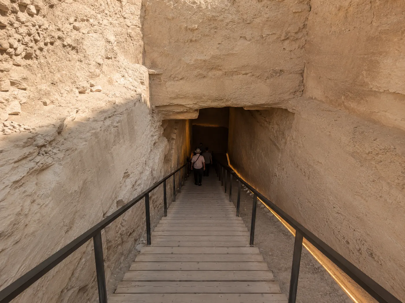 A wooden walkway with handrails dropping into a plain rock-cut opening in the hillside — how every tomb in the valley begins